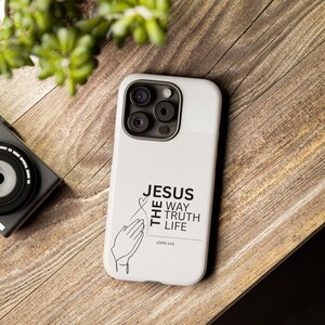 Könnte beinhalten: Eine weiße Smartphone-Hülle mit einer schwarzen Strichzeichnung von betenden Händen und einem kleinen Herz darüber. Der Text JESUS THE WAY TRUTH LIFE sowie JOHN 14:6 ist in einer klaren, schwarzen serifenlosen Schrift auf der Rückseite abgebildet.