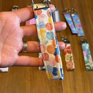 May include: A fabric wristlet keychain featuring a white background with a pattern of overlapping orange, yellow, and blue watercolor-style circles. The strap is secured with a silver-toned metal hardware clamp and a circular split ring at the top.
