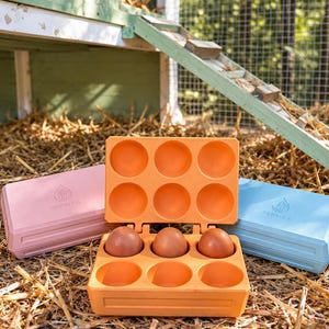 Pode incluir: Três caixas de ovos retangulares em rosa pastel, laranja brilhante e azul claro. A caixa laranja está aberta, revelando seis compartimentos circulares, com três ovos castanhos na fila inferior. Cada tampa apresenta o texto PERMEA.