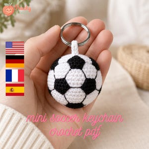 May include: A small, spherical football keychain crafted from white and black yarn using a crochet technique. The ball features the classic pentagonal and hexagonal pattern and is attached to a circular silver-toned metal split ring.