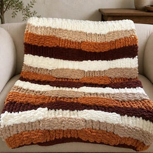 May include: A chunky knit throw blanket featuring horizontal stripes in cream, beige, burnt orange, and dark brown. The blanket is crafted from thick, soft yarn with a textured, looped appearance, creating a layered and cozy look across the fabric.