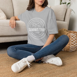 Peut inclure: Un t-shirt gris chiné à manches courtes avec un motif circulaire blanc sur la poitrine. Le design comprend une chaîne de montagnes, un soleil et le texte : MAKING MEMORIES TOGETHER, SUMMIT TRIP, 2026, COLORADO, USA, dans un cercle à double ligne.
