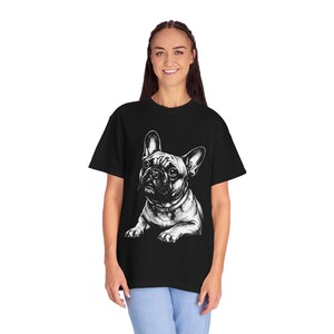 Puede incluir: Una camiseta negra de manga corta con una detallada ilustración gráfica en blanco de un bulldog francés en posición sentada. El perro aparece con orejas grandes y un collar, centrado en la parte delantera de la prenda, que se lleva con pantalones vaqueros azules.