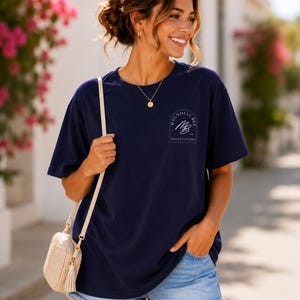 May include: A navy blue short-sleeved t-shirt featuring a white arched logo on the chest that reads Magnolia Bee with the initials MB. The shirt is paired with light blue denim shorts with a frayed hem and a small beige woven crossbody bag.