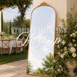 Puede incluir: Un espejo de pie alto y arqueado con un marco dorado fino y un adorno decorativo en la parte superior. La superficie del espejo refleja el cielo y los árboles. El texto "WEDDING MIRROR SIGN Mockup" está superpuesto en blanco sobre el espejo.