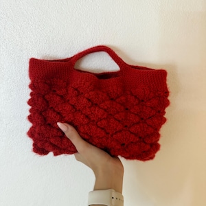 Peut inclure: Un sac à main en crochet rouge vif présentant un motif texturé en points noisette sur tout le corps. Le sac a une forme rectangulaire avec deux anses intégrées, fabriqué à partir d'un matériau en fil doux et duveteux d'une couleur rouge unie.