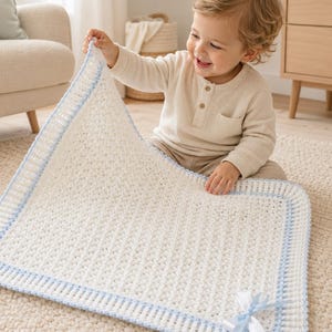 Può includere: Una coperta rettangolare all'uncinetto di colore bianco con un motivo a trama goffrata. La coperta è rifinita con un bordo a coste in filato azzurro e bianco ed è decorata con un piccolo fiocco in nastro di raso azzurro e bianco.