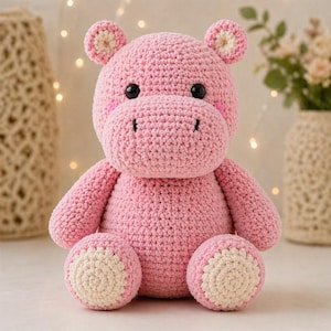 May include: A pink crocheted hippopotamus stuffed animal with a rounded body, large head, and small ears. It features black plastic eyes, a stitched mouth, and cream-colored circular patches on the soles of its feet and the center of its ears.