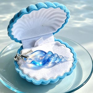 May include: A spiral-shaped glass pendant with blue and clear iridescent tones, attached to a silver-colored chain. The pendant rests inside a light blue, velvet-lined, shell-shaped jewelry box that is open to display the item clearly.