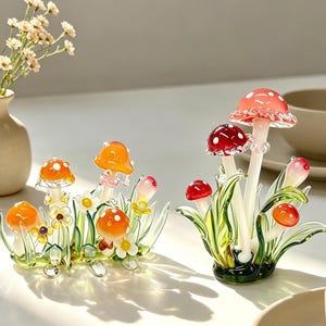 May include: Two glass mushroom sculptures. The left piece features orange caps with white spots and small yellow flowers amidst green glass blades. The right piece displays red caps with white spots on white stems, surrounded by green glass foliage.