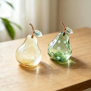 May include: Two pear-shaped glass ornaments. One is translucent yellow, and the other is translucent green with white polka dots. Each features a metallic copper-coloured stem and a single small, light blue glass leaf attached to the top.