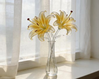 May include: Two yellow glass lily flowers with delicate petals and dark red stamens, featuring small clear droplets on the surface. The flowers are arranged in a slender, transparent glass vase with a narrow neck and a flared base.