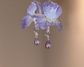 May include: A pair of dangle earrings featuring translucent purple and white flower-shaped charms with gold-colored accents at the center. Each earring hangs from a silver-toned metal hook and includes a small, teardrop-shaped purple bead.