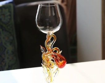 May include: A clear glass goblet featuring a stem designed with swirling, flame-like glass elements in shades of yellow, orange, and deep red. The base is circular and transparent, supporting the intricate, multi-colored glass stem.