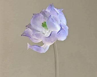 May include: A handcrafted glass flower sculpture featuring delicate, translucent light purple petals that transition to a clear base. The center of the bloom displays a vibrant green stamen, and the entire structure is supported by a clear glass stem.