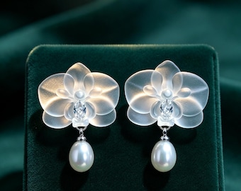 May include: A pair of earrings featuring translucent, frosted orchid-shaped petals with a central marquise-cut clear stone. Each earring is finished with a single, smooth, white teardrop-shaped pearl suspended from the base of the flower design.