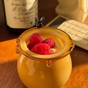 May include: A round, amber-colored glass jar with a thick, rounded rim and a small, dark blue glass bee figurine attached to the top edge. The jar is filled with a creamy white substance topped with several fresh, bright red raspberries.