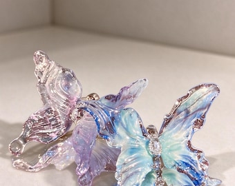 May include: Two butterfly-shaped hair clips featuring translucent, iridescent wings in shades of blue, purple, and teal. Each clip has a metallic body accented with small, clear, sparkling stones along the center, creating a shimmering effect.