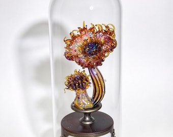 May include: A glass sculpture of two sea anemones with orange, purple, and yellow tentacles inside a clear glass dome. The sculpture sits on a dark wooden base with a metal pedestal and a small metal plaque featuring the text Actinia.