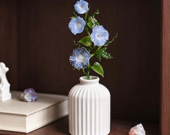 May include: A white ceramic vase with vertical ribbing holds a glass sculpture of morning glory flowers. The flowers are light blue with white centers, attached to green glass stems and leaves that feature delicate, curling tendrils.