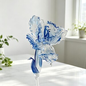 Peut inclure: Une sculpture de fleur en verre fabriquée à la main, dotée de pétales transparents aux bords bleu vif et de détails bleus complexes au centre. La fleur est fixée sur une tige en verre clair et élancée avec une feuille bleue en guise d'accent.