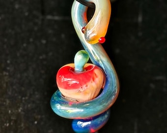 May include: A glass pendant featuring a coiled snake with a translucent blue and orange body and small red eyes, wrapped around a central red and white apple-shaped glass element, suspended from a thin black cord.