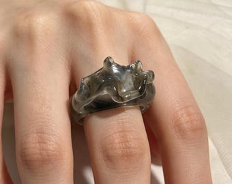 May include: A translucent ring featuring a swirling, marbled pattern of smoky gray and white tones. The band has an irregular, wavy, and organic shape with raised, rounded protrusions along the top edge, creating a fluid, sculptural appearance.