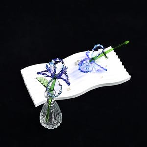 May include: Two blue glass iris flowers with green stems. One flower is upright in a clear, ribbed glass vase, while the other lies horizontally on a white, rectangular, wavy-edged tray. The petals feature a translucent, textured blue finish.
