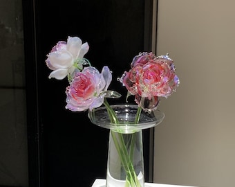 May include: Three glass flowers with iridescent petals in shades of pink, white, and translucent clear, featuring delicate ruffled edges. The flowers are arranged in a clear, cylindrical glass vase with a wide, flat rim at the top.