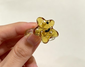 May include: A clear ring featuring a translucent amber-colored star-shaped accent. The star has a smooth, glossy finish and is decorated with two small, black, raised dots representing eyes and a curved black line forming a simple smiling mouth.