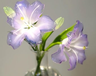 May include: Two translucent purple lily flowers with white stamens and yellow centers, featuring green stems and buds, arranged in a clear glass vase. The petals have a delicate, glossy finish with light purple edges and lighter centers.