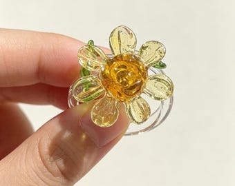 May include: A clear glass ring featuring a central flower design with a deep amber-colored core and translucent pale yellow petals. The flower is accented with small green glass leaves at the base, all set upon a simple, transparent circular band.