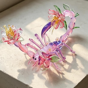 May include: A glass sculpture of a scorpion with a translucent pink and purple body, featuring delicate glass flowers in shades of pink, yellow, and green attached to its tail and back, with small black eyes and slender, segmented legs.