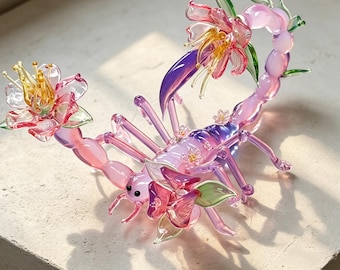 May include: A glass sculpture of a scorpion with a translucent pink and purple body, featuring delicate glass flowers in shades of pink, yellow, and green attached to its tail and back, with small black eyes and slender, segmented legs.