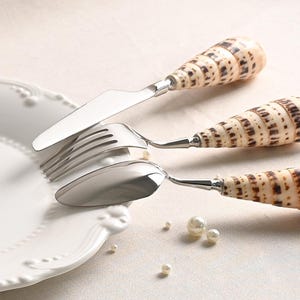 May include: A set of three stainless steel utensils including a knife, fork, and spoon, each featuring a handle crafted from a natural cone-shaped seashell with brown and cream patterns, resting on the edge of a white ceramic plate.