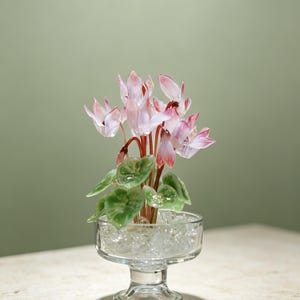 May include: A glass sculpture of a cyclamen plant with delicate pink and white petals and green leaves. The plant is set in a clear glass pedestal bowl filled with small, translucent glass beads that mimic the appearance of water or soil.