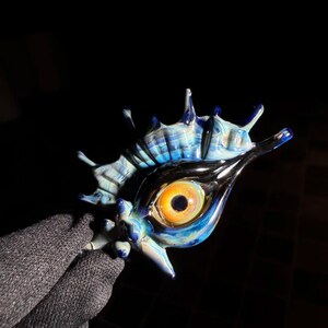 May include: A glass sculpture shaped like a spiky seashell featuring a central realistic orange and black eye. The shell has swirling patterns of blue, white, and black glass, with sharp, pointed protrusions extending from the main body.