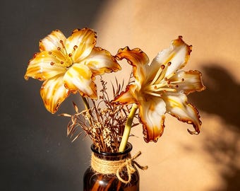 May include: Two glass lily flowers with white and amber-colored petals featuring ruffled edges and prominent stamens. The flowers are arranged in a small, dark brown glass bottle wrapped with a piece of natural jute twine around the neck.