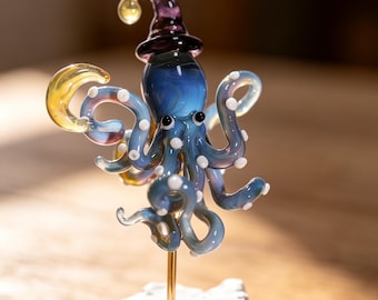 May include: A glass octopus figurine in shades of blue with white spots, wearing a purple pointed hat. The octopus holds a yellow crescent moon and is mounted on a thin gold-colored rod attached to a textured white rock-like base.