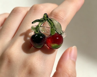 May include: A clear glass ring featuring a decorative cherry motif. The design includes two small, rounded glass cherries, one in a deep red and one in a dark green, attached to a clear glass band with two green glass leaves on top.