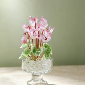 May include: A glass sculpture of a cyclamen plant with delicate pink and white petals, thin brown stems, and green heart-shaped leaves. The plant is set in a clear glass pedestal bowl filled with clear glass shards that mimic ice or water.