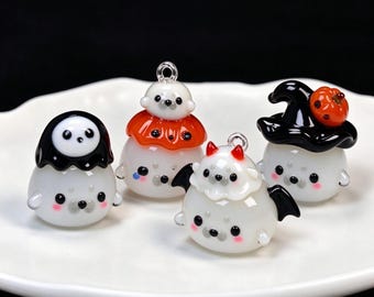 May include: Four small, round, white glass seal charms with black eyes and pink cheeks. One wears a black ghost hood, one has a small seal on an orange collar, one has black bat wings and red horns, and one wears a black witch hat with a pumpkin.