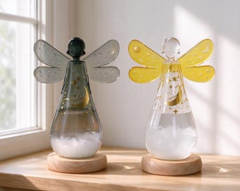 May include: Two glass weather storm glass ornaments shaped like angels with wings. One is dark gray and the other is yellow. Both feature gold celestial patterns of moons and stars on the glass body and sit on round light wood bases.