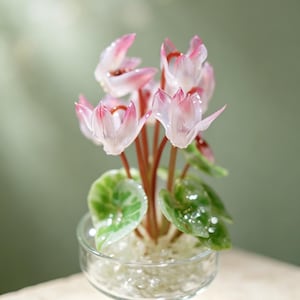 May include: A glass sculpture of a cyclamen plant with delicate, translucent pink and white petals, slender reddish-brown stems, and variegated green leaves, all contained within a clear glass pedestal bowl filled with small, clear glass beads.