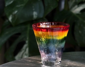 May include: A glass tumbler featuring a vibrant, multi-colored gradient design that transitions from a deep red rim through orange, yellow, green, blue, and purple at the base. The glass has a textured, rippled surface and a flared shape.