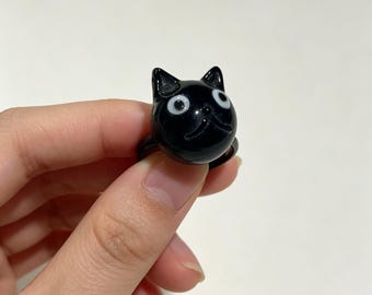 May include: A black ring featuring a glossy, spherical cat head design. The cat has two pointed ears, a small nose, and two wide, white circular eyes with black pupils, creating a distinct and recognizable feline facial expression.