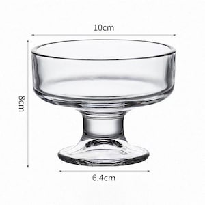 May include: A clear glass dessert bowl with a wide circular rim and a sturdy pedestal base. The bowl measures 10 cm in diameter at the top, 6.4 cm at the base, and stands 8 cm tall. The design features a smooth, transparent surface throughout.