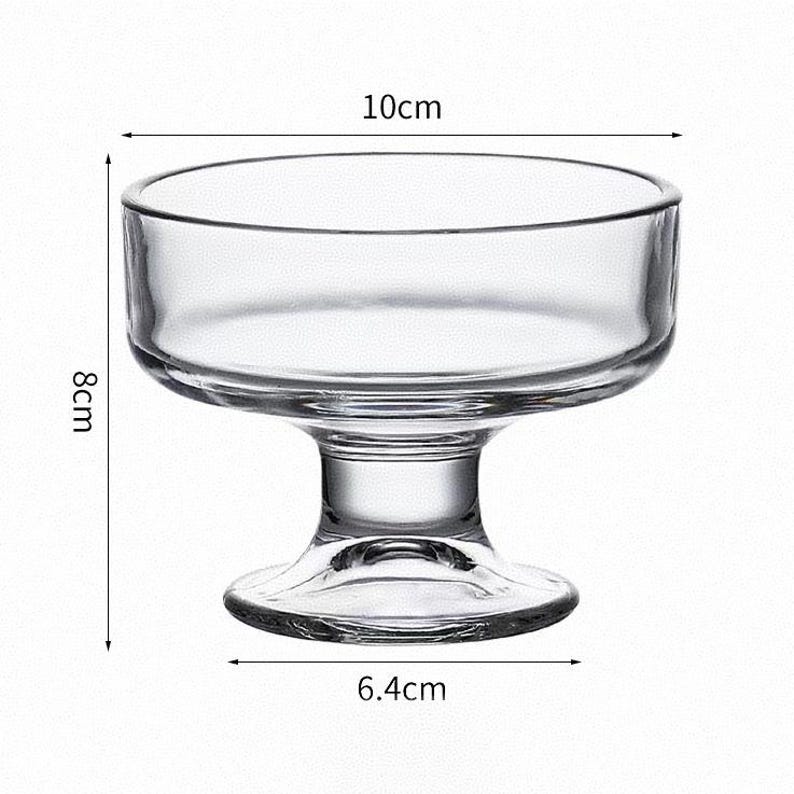 May include: A clear glass dessert bowl with a wide circular rim and a sturdy pedestal base. The bowl measures 10 cm in diameter at the top, 6.4 cm at the base, and stands 8 cm tall. The design features a smooth, transparent surface throughout.
