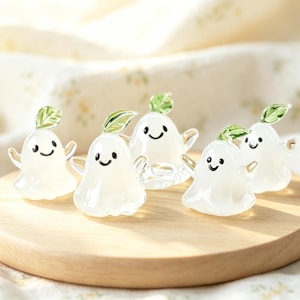 Pode incluir: Cinco pequenas figuras de fantasmas em vidro branco translúcido, cada uma com um rosto sorridente e uma folha verde no topo. Os fantasmas têm corpos arredondados e pequenos braços estendidos, dispostos sobre uma base circular de madeira.