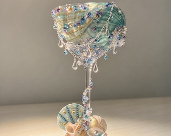 May include: A decorative goblet featuring a large seashell bowl adorned with blue, purple, and clear beads and silver chains with teardrop charms. The stem is wrapped in colorful beads, and the base is decorated with various seashells and beads.